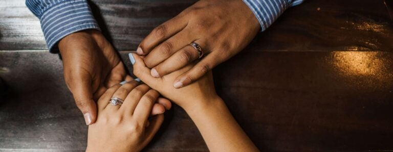 Four hands with wedding rings holding each other's hands protectively.