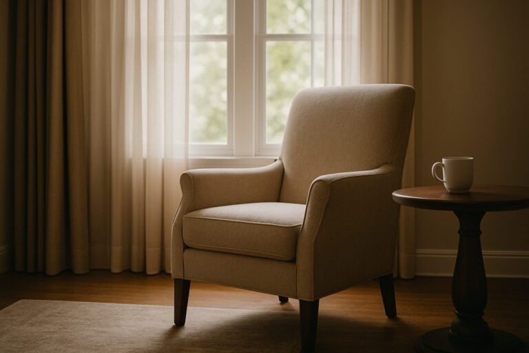 Upholstered chair with a single cup on a side table with two windows with curtains behind it representing a person alone hoping to improve their marriage.