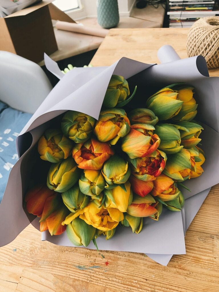 A bouquet of colorful tulips wrapped in paper, set on a wooden table indoors.