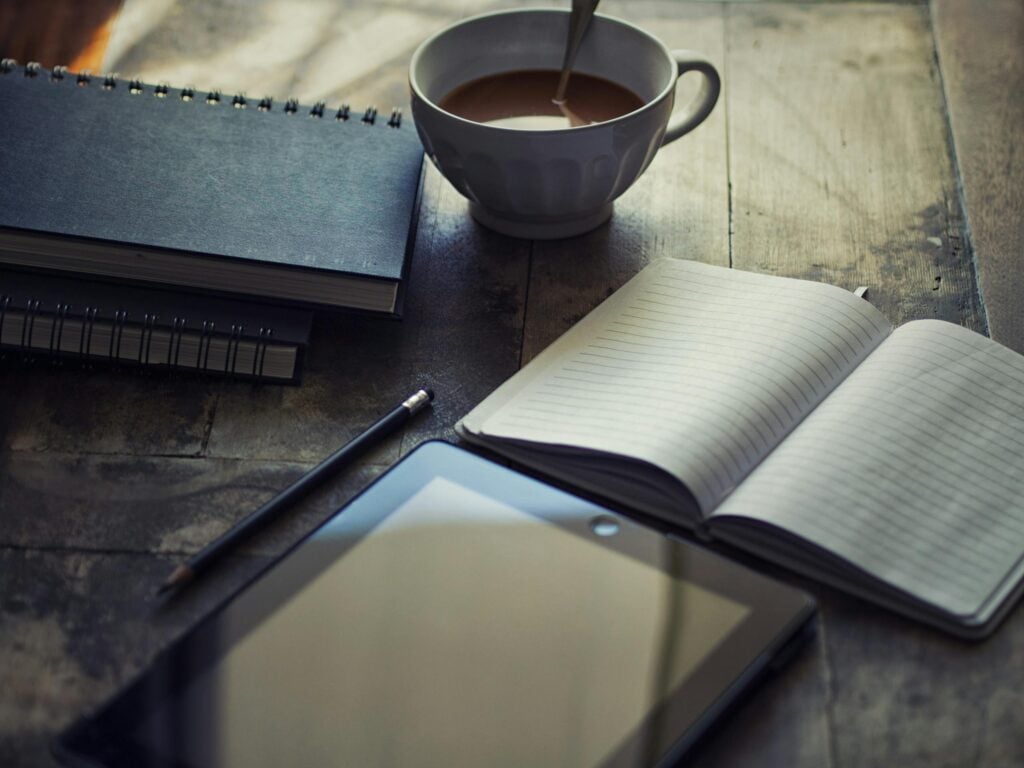 A cozy workspace setup with coffee, notebook, and tablet on a wooden table.