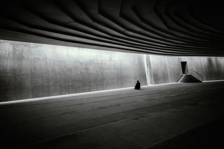 A lone person meditates in a vast, modern, minimalist indoor hall with dramatic architecture.