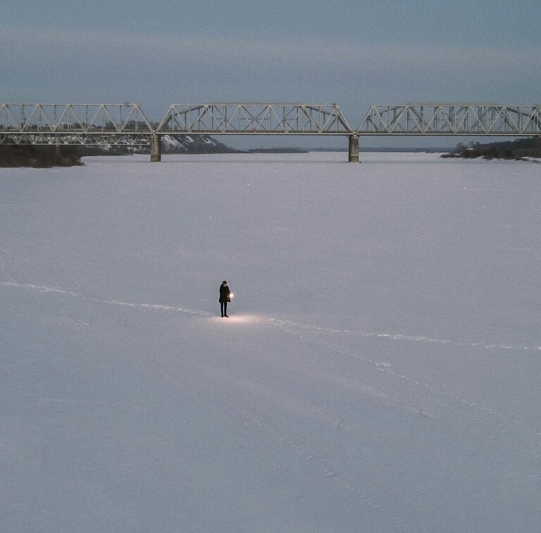 A lone person walks across a snow-covered frozen river, carrying a lantern, beneath a distant bridge.