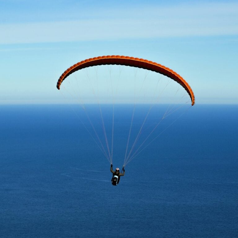 A person paragliding with an orange chute over a vast ocean under clear blue skies.