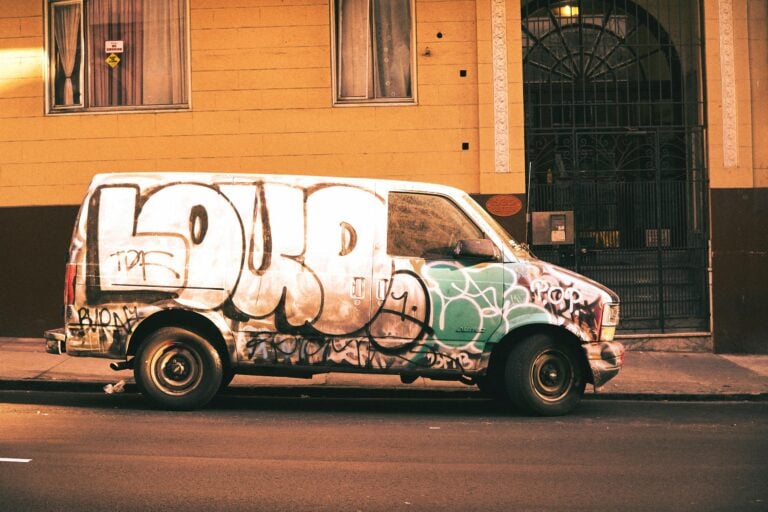 Urban street scene featuring a graffiti-covered van parked near a building with an arched doorway.